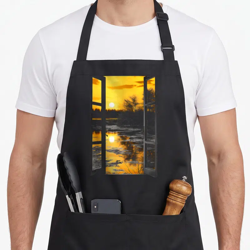 Tablier de cuisine noir avec illustration d'une fenêtre ouverte sur un paysage au coucher de soleil orange et gris.