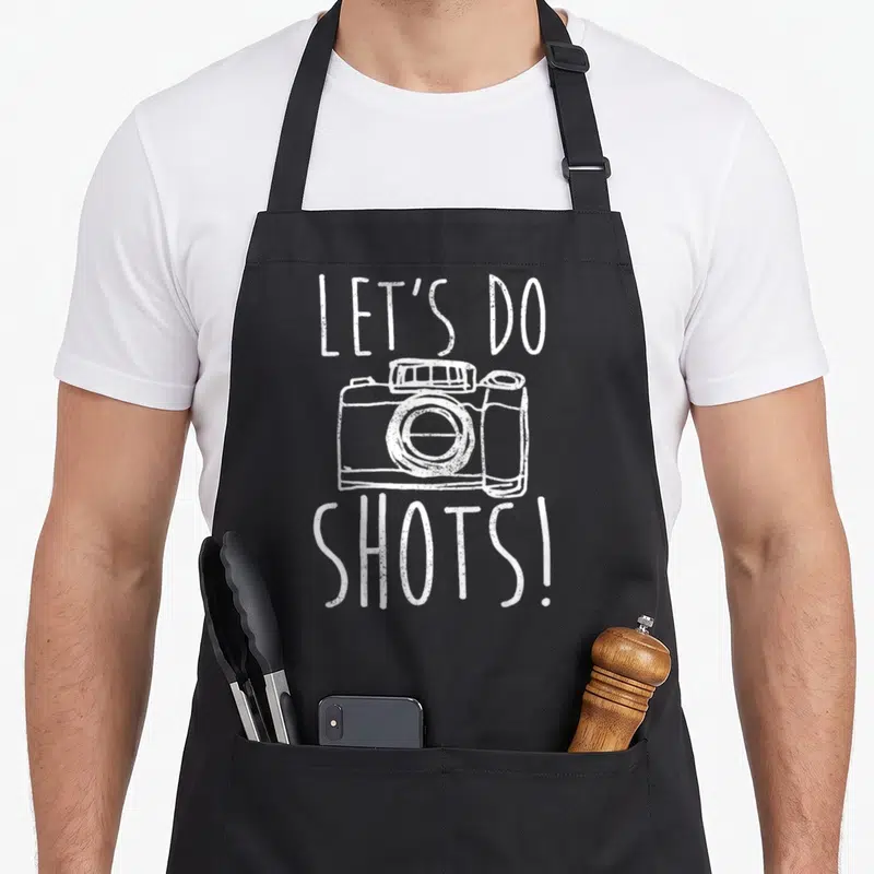 Tablier de cuisine noir avec inscription Let's do shots et illustration de caméra photographique en blanc.
