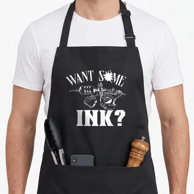 Tablier de cuisine noir avec une machine à tatouer, un pichet d'encre et le texte Want some ink en blanc.