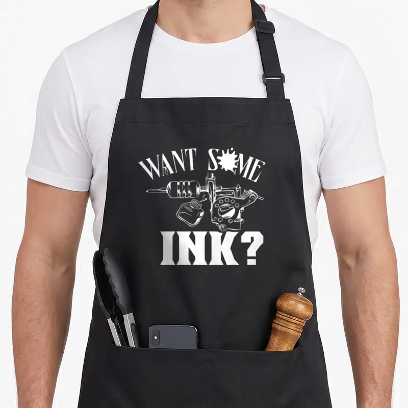 Tablier de cuisine noir avec une machine à tatouer, un pichet d'encre et le texte Want some ink en blanc.