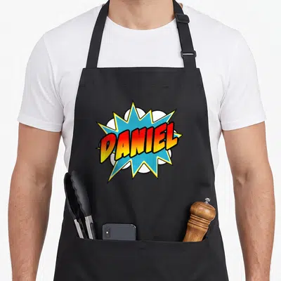 Tablier de cuisine noir avec une explosion bleue portant le nom Daniel en lettres rouges et jaunes au centre.