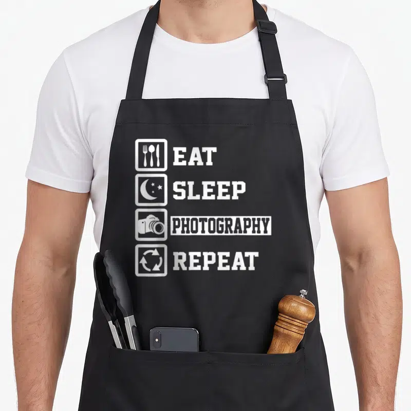 Tablier de cuisine noir avec les mots Eat Sleep Photography Repeat et quatre icônes blanches alignées verticalement.