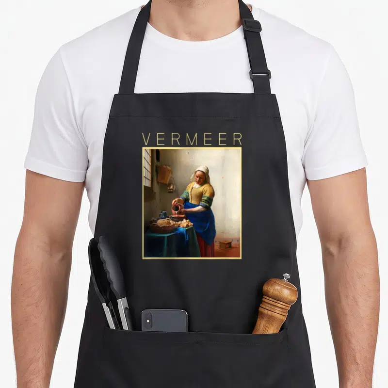 Tablier de cuisine noir avec reproduction d'une femme versant du lait et le nom Vermeer en lettres dorées au-dessus.