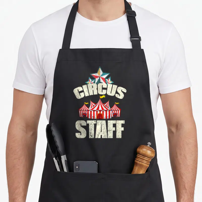 Tablier de cuisine noir avec illustration de tente de cirque et texte Circus Staff en blanc vieilli.
