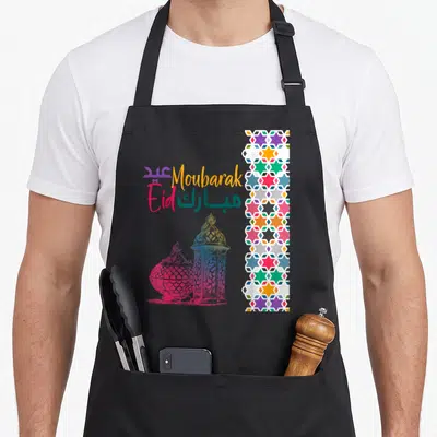 Tablier de cuisine noir avec inscription « Eid Moubarak », lanternes colorées et frise de motifs géométriques arabesques sur le côté droit.