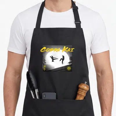 Tablier de cuisine noir avec deux silhouettes martiales sautant devant une lune et le texte Cobra Kai en jaune.