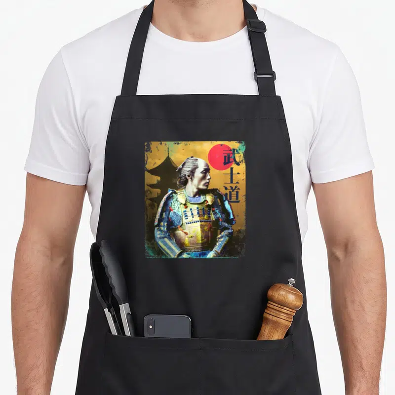 Tablier de cuisine noir avec illustration d'un samouraï vintage, texte et soleil rouge sur fond doré texturé.