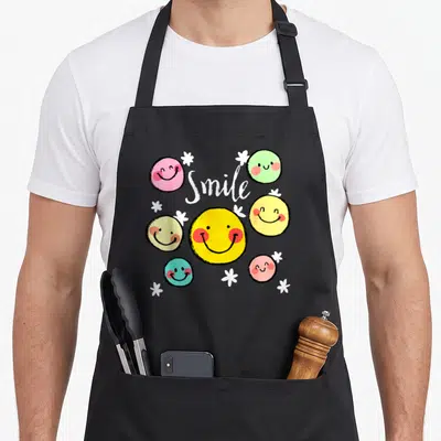 Tablier de cuisine noir avec des visages souriants colorés, des étoiles blanches et le mot Smile en écriture cursive au centre.