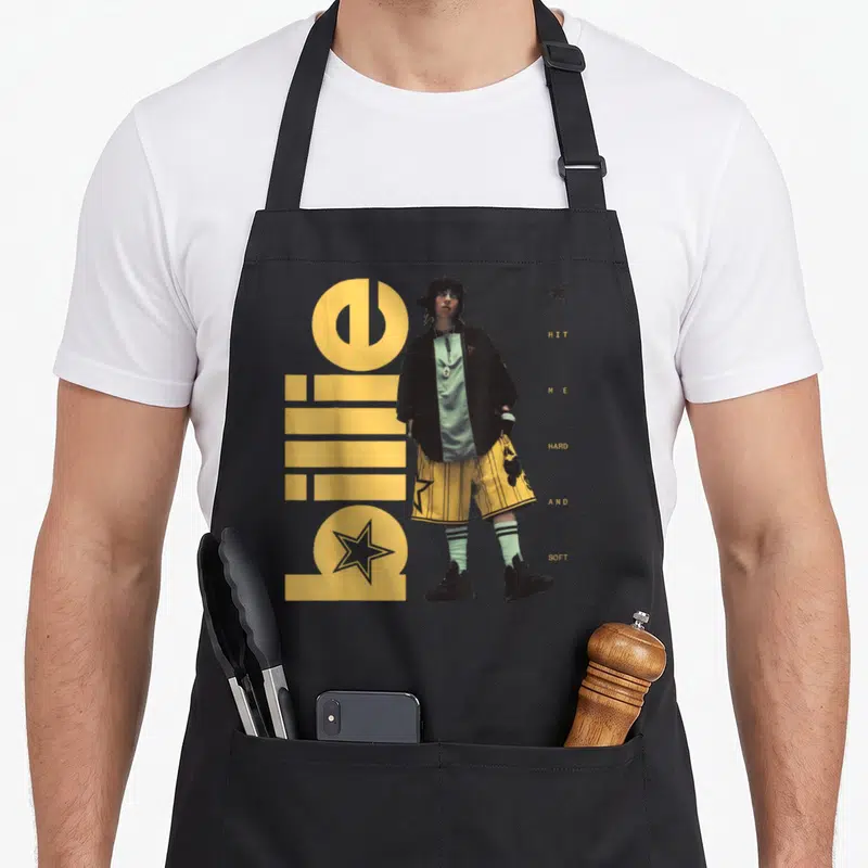 Tablier de cuisine noir avec une illustration de Billie Eilish debout, le texte Hit Me Hard and Soft et des étoiles jaunes.