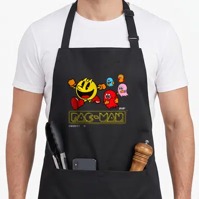 Tablier de cuisine noir avec Pac-Man jaune courant suivi de quatre fantômes colorés et le texte 2UP Credit.
