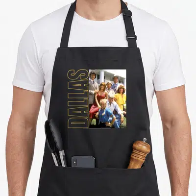 Tablier de cuisine noir avec photo de famille des Ewing et texte Dallas vertical doré sur fond gris uni.