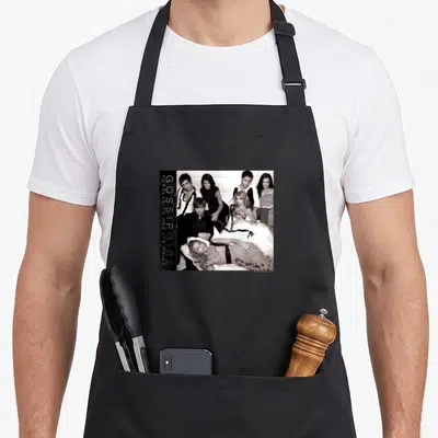 Tablier de cuisine noir avec une illustration groupante en noir et blanc et le texte Gossip Girl Populaire a un charme sur le côté gauche.