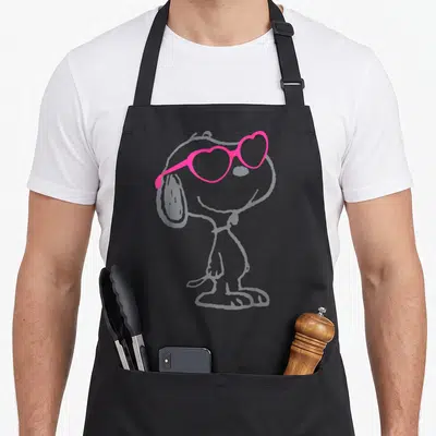 Tablier de cuisine noir avec un croquis gris de Snoopy coiffé de lunettes de soleil roses en forme de cœurs.