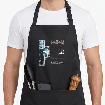 Tablier de cuisine noir avec illustration d'un appareil photo argenté et le texte Def Leppard Photograph en rose et blanc.