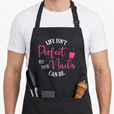 Tablier de cuisine noir avec illustration vernis à ongles rose et texte Life Isn't Perfect But Your Nails Can Be en blanc et rose.