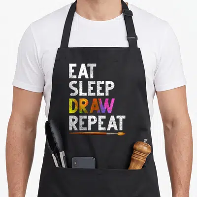Tablier de cuisine noir avec inscription « EAT SLEEP DRAW REPEAT » et un pinceau peint en bois sous le texte.