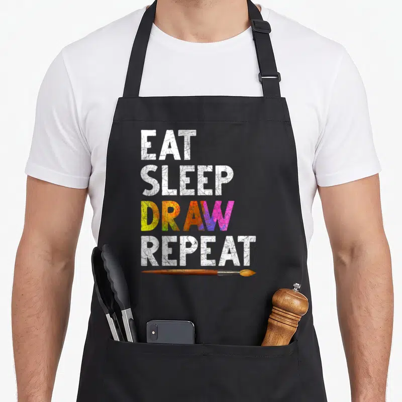 Tablier de cuisine noir avec inscription « EAT SLEEP DRAW REPEAT » et un pinceau peint en bois sous le texte.