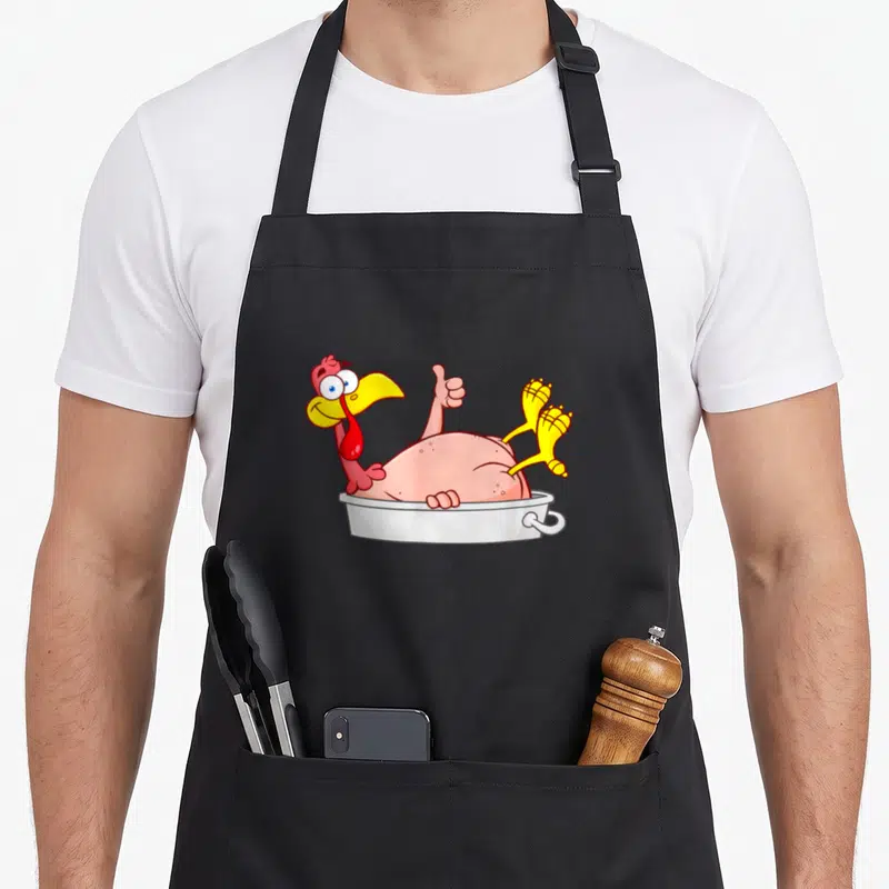 Tablier de cuisine noir avec une illustration colorée d'une dinde de dessin animé dans un plat de cuisson, faisant un geste amical.