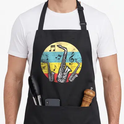 Tablier de cuisine noir avec illustration circulaire de trois saxophones et notes de musique sur fond rayé jaune bleu rouge.