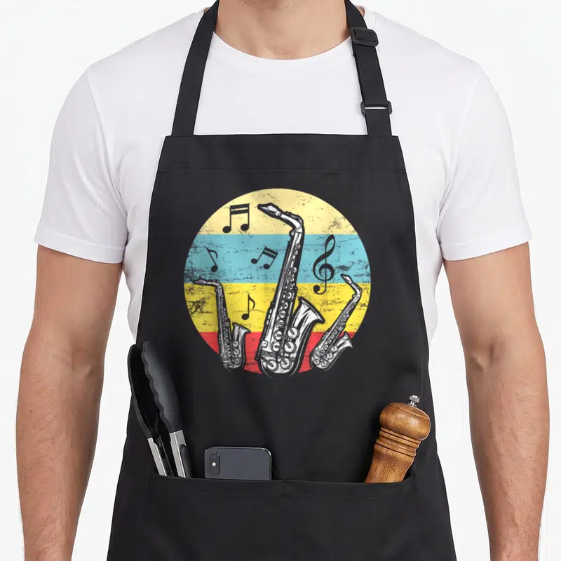 Tablier de cuisine noir avec illustration circulaire de trois saxophones et notes de musique sur fond rayé jaune bleu rouge.