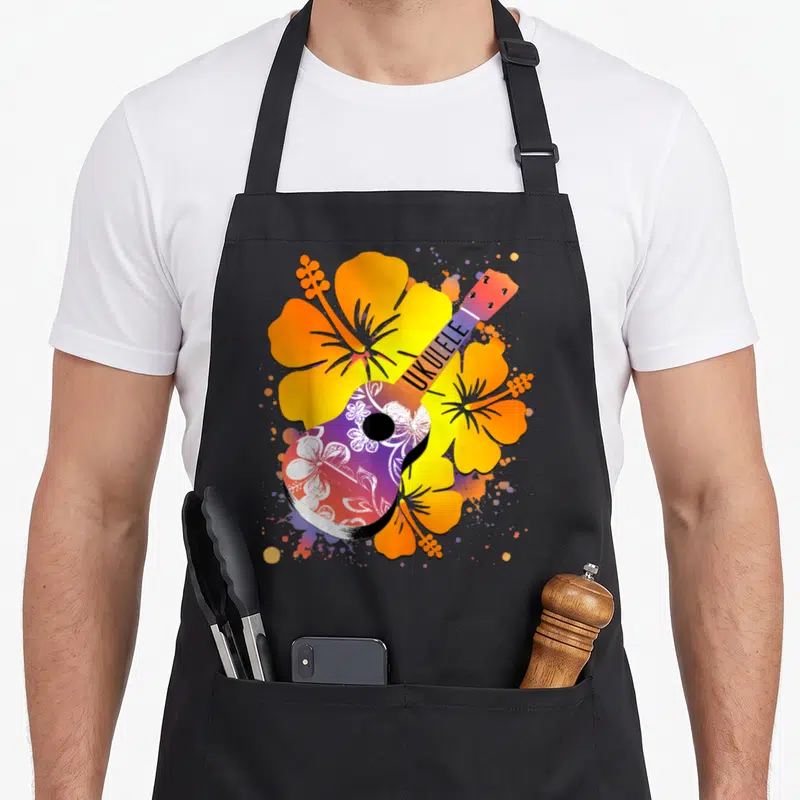 Tablier de cuisine noir avec ukulélé décoré de motifs floraux blancs et entouré de grandes fleurs d'hibiscus orange et jaunes.