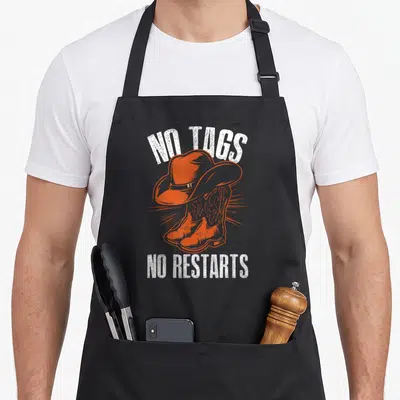 Tablier de cuisine noir avec illustration orange de chapeau de cowboy et bottes, texte blanc No Tags No Restarts.