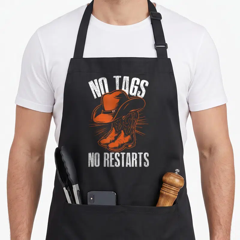 Tablier de cuisine noir avec illustration orange de chapeau de cowboy et bottes, texte blanc No Tags No Restarts.
