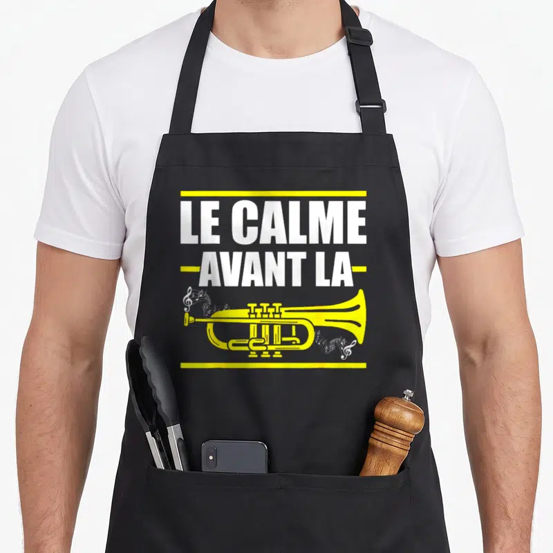 Tablier de cuisine noir avec illustration d'une trompette jaune et texte blanc Le calme avant la trompette encadré de lignes jaunes.