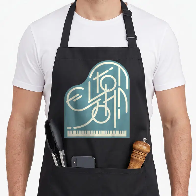 Tablier de cuisine noir avec logo piano Elton John stylisé en lettres blanches sur fond bleu sarcelle et clavier en bas.