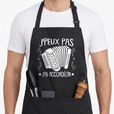 Tablier de cuisine noir avec illustration blanche d'accordéon et le texte Jpeux pas j'ai accordéon encadré de volutes décoratives.