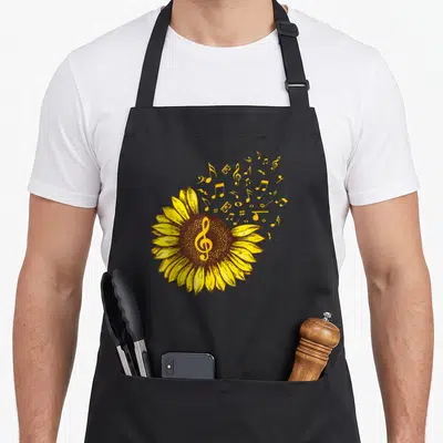Tablier de cuisine noir avec une illustration de tournesol jaune et de notes de musique dorées s'échappant du centre floral.
