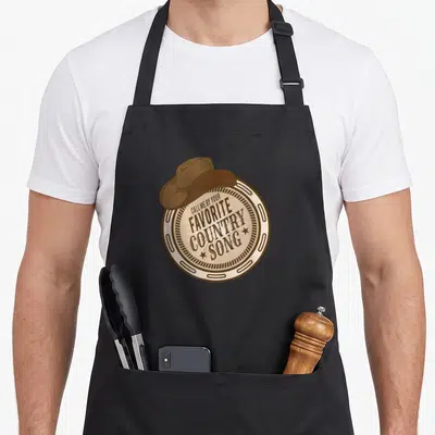 Tablier de cuisine noir avec une illustration circulaire beige et marron représentant un chapeau cowboy sur le texte Call Me By Your Favorite Country Song.