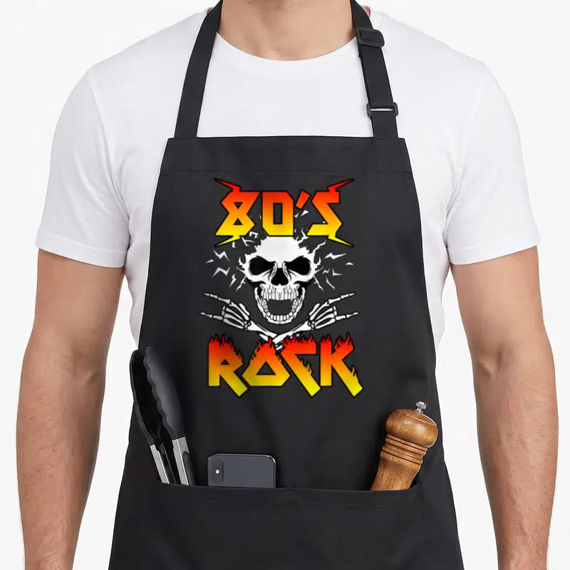Tablier de cuisine noir avec un crâne blanc, des os croisés et le texte 80's Rock en lettres flamboyantes orange et jaunes.