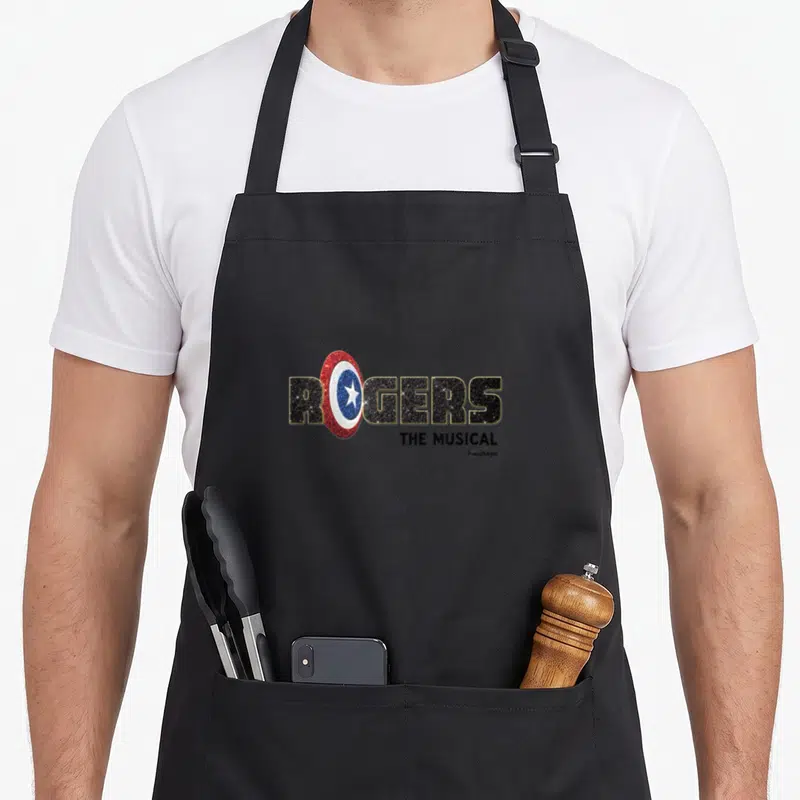Tablier de cuisine noir avec le logo Rogers The Musical intégrant un bouclier rouge pailleté au centre du texte.
