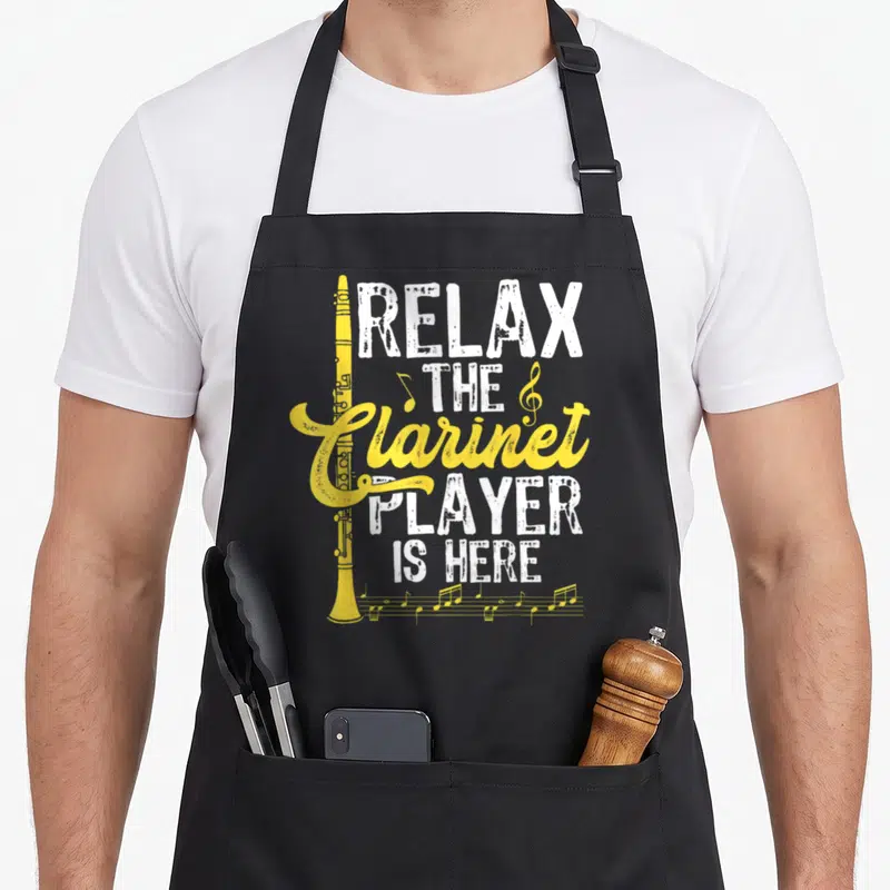 Tablier de cuisine noir avec une clarinette jaune et le texte Relax The Clarinet Player Is Here imprimé en blanc et jaune.
