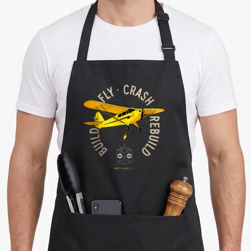 Tablier de cuisine noir avec illustration d'un avion RC jaune, une télécommande et le texte Fly Crash Rebuild.