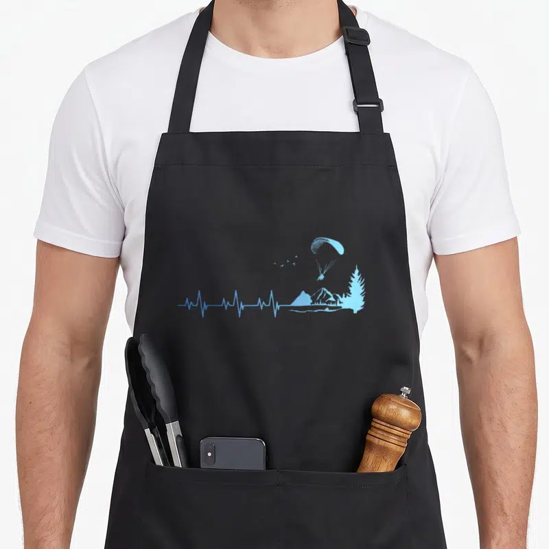Tablier de cuisine noir avec un parapente bleu, une montagne et un arbre stylisés sur fond uni.