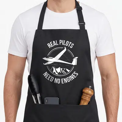 Tablier de cuisine noir avec l'inscription Real Pilots Need No Engines et une illustration de planeur au-dessus de montagnes.