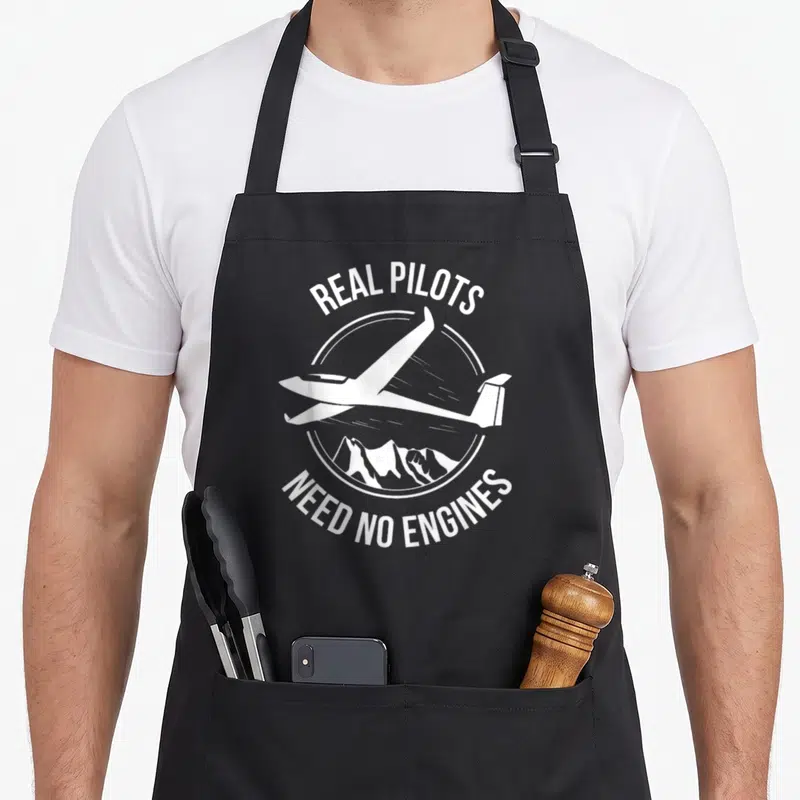 Tablier de cuisine noir avec l'inscription Real Pilots Need No Engines et une illustration de planeur au-dessus de montagnes.
