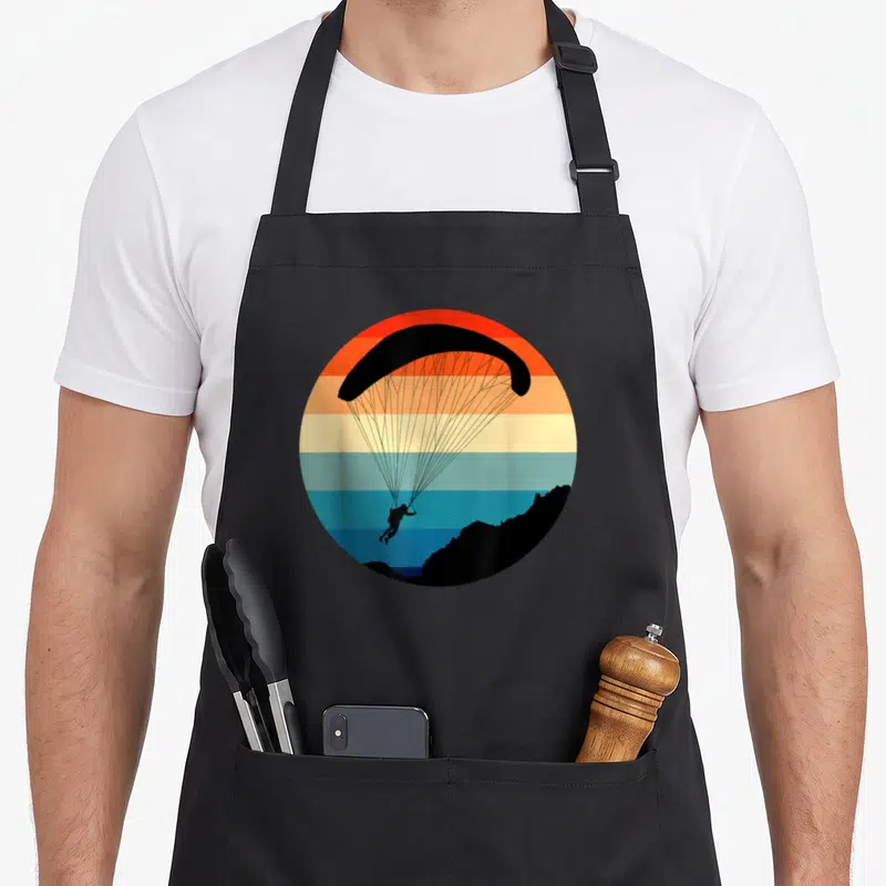 Tablier de cuisine noir avec un cercle coloré représentant un parapente et un pilote au-dessus d'une montagne.