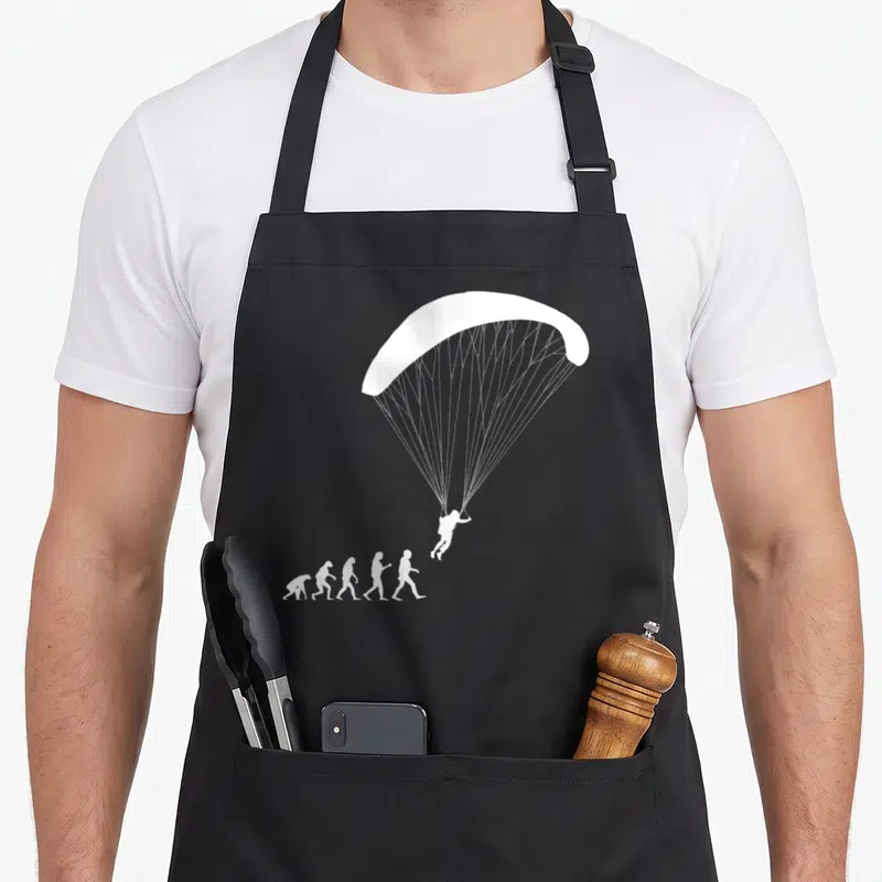 Tablier de cuisine noir avec silhouette blanche représentant l'évolution humaine se terminant par un parapentiste en vol.