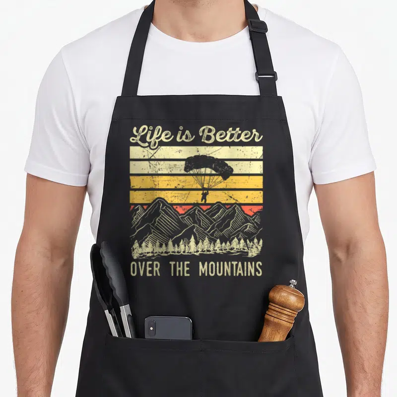 Tablier de cuisine noir avec une illustration vintage d'un parapentiste au-dessus de montagnes et de forêts sous un ciel strié.