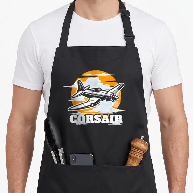 Tablier de cuisine noir avec illustration d'un avion Corsair blanc sur fond de soleil orange et nuages.