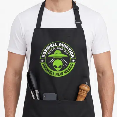 Tablier de cuisine noir avec un logo circulaire vert montrant un engin volant et une tête d'alien aux grands yeux noirs.