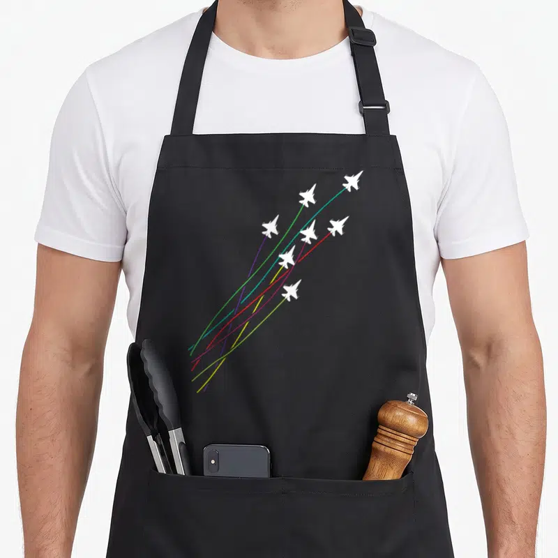 Tablier de cuisine noir avec avions de chasse blancs laissant des traînées multicolores en ascension diagonale.