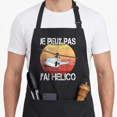 Tablier de cuisine noir avec inscription Je peux pas j'ai helico et illustration d'hélicoptère sur fond circulaire dégradé orange et rouge.