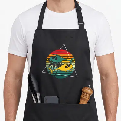 Tablier de cuisine noir avec hélicoptères rétro, soleil strié et palmiers dans un triangle blanc.