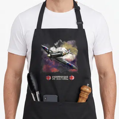 Tablier de cuisine noir avec illustration d'un avion Supermarine Spitfire volant dans des nuages colorés et le texte Spitfire encadré de drapeaux britanniques.