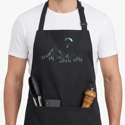 Tablier de cuisine noir avec illustration bleue d'un parapente au-dessus de montagnes et d'arbres conifères stylisés.
