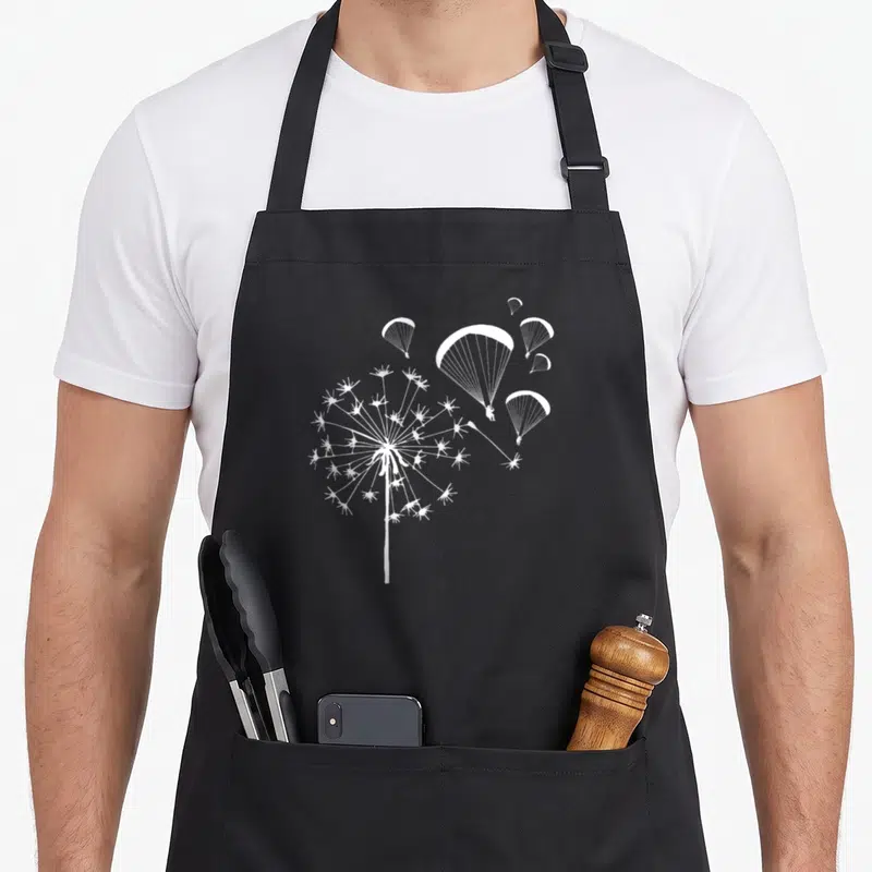 Tablier de cuisine noir avec illustration blanche d'un dandelion dont les graines deviennent des parachutes de parapente.